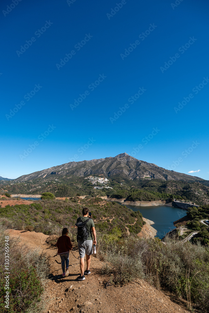 Father and daughter hiking on mountain trail near scenic lake under clear blue sky, La Concepcion reservoir in Marbella Father and daughter hiking on mountain trail near scenic lake under clear blue sky, La Concepcion reservoir in Marbella