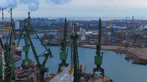 Slow aerial orbit around green cranes, coal plant EC 2 smokestacks in distance, and industrial zones at Gdansk Shipyard along river