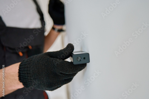 Construction worker in gloves uses laser distance measurer to inspect wall. Builder using digital tool for precision measurement during repair work. Construction and renovation concept