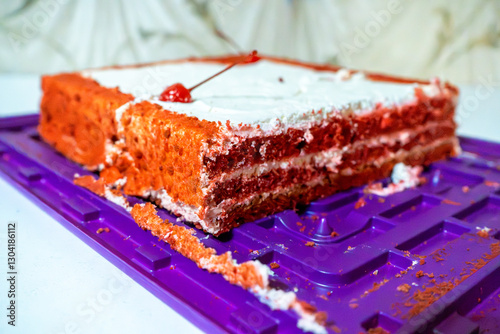 A close-up of a red velvet cake with white frosting and a cherry on top, sitting on a purple tray. The layers of the cake are visible, showcasing the rich red color contrasting with the white cream
