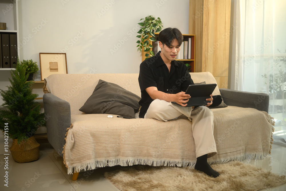 Obraz premium Relaxed asian man sitting on cozy sofa in a modern living room and using digital tablet