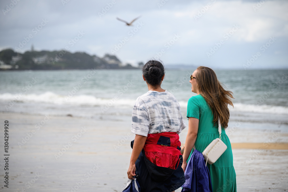 Two women chatting and walking at Takapuna Beach. Out-of-focus seagull flying in the sky. Auckland.