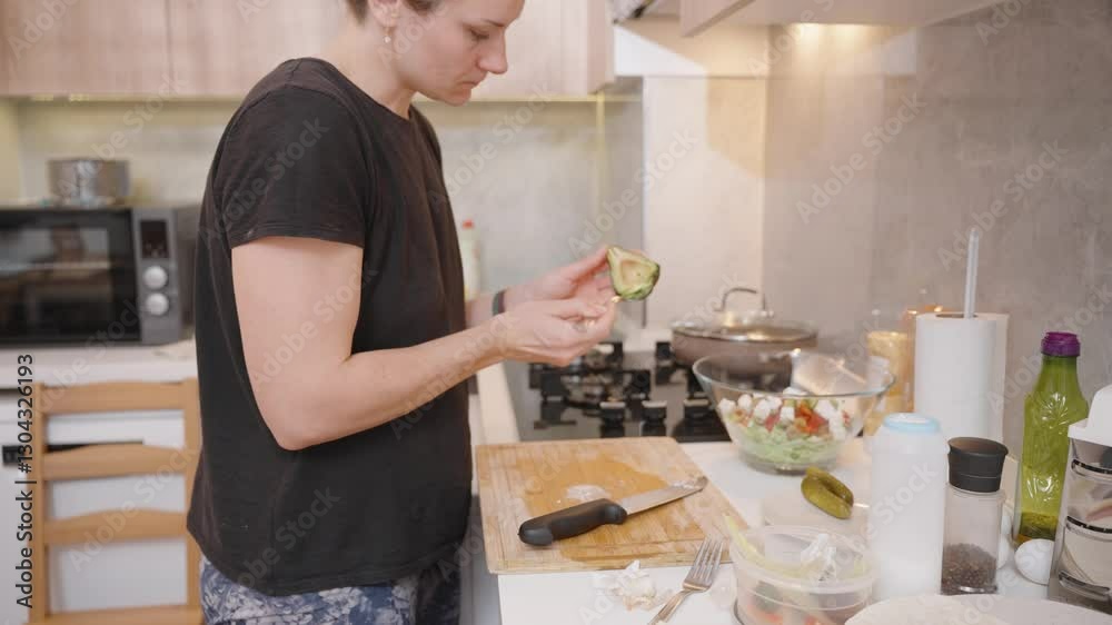 Woman Scooping Out an Avocado with a Spoon in a Bright Kitchen, Preparing a Fresh Salad with a Glass Bowl Full of Ingredients on the Counter Woman Scooping Out an Avocado with a Spoon in a Bright Kitchen, Preparing a Fresh Salad with a Glass Bowl Full of Ingredients on the Counter