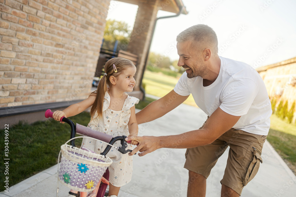Naklejka premium Portrait of a smiling father helping his little girl to learn how to ride a bicycle in backyard.