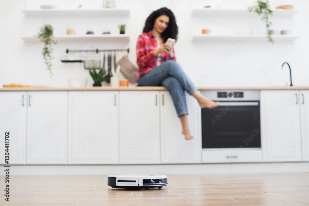 Positive atmosphere modern kitchen African woman sitting counter using smartphone autonomous robot vacuum cleaner operating visible interaction technology convenience lifestyle comfort clean space