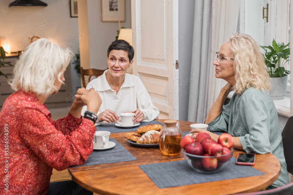 Three elderly women sitting at table chatting joyfully. Elegance blonde senior women chill together talking. Female aged friends having tea time at home and communicating smiling Three elderly women sitting at table chatting joyfully. Elegance blonde senior women chill together talking. Female aged friends having tea time at home and communicating smiling