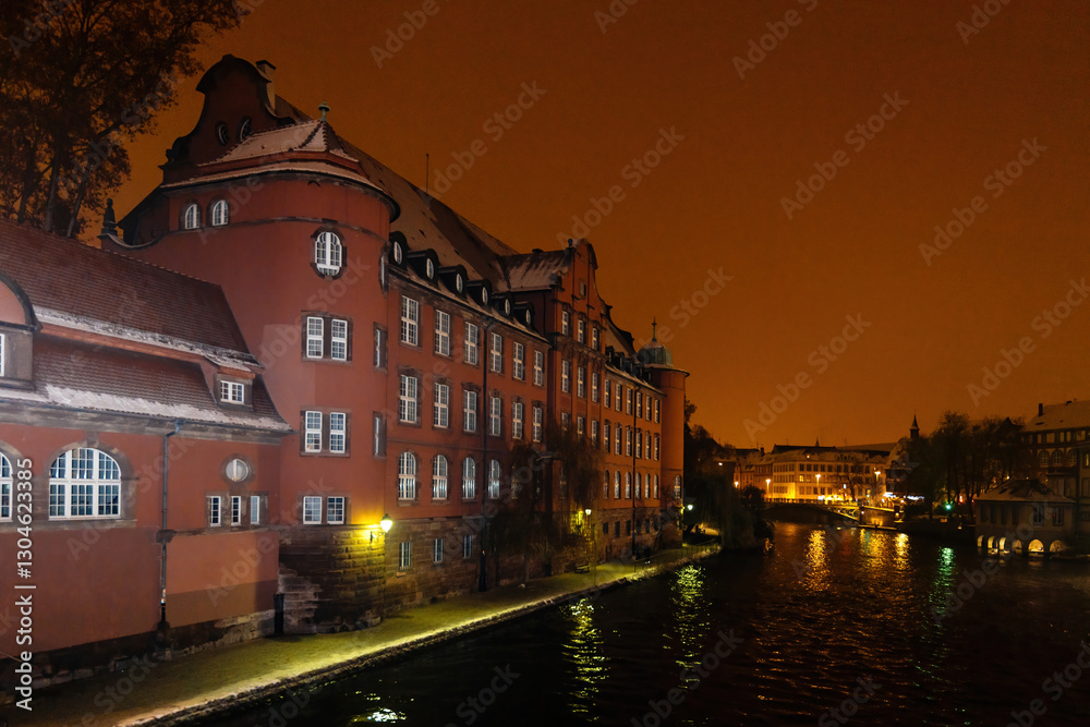 Fototapeta premium Orange hues envelop the historic Saint Thomas school building along the Ill River, viewed from Pont Saint-Martin at night.
