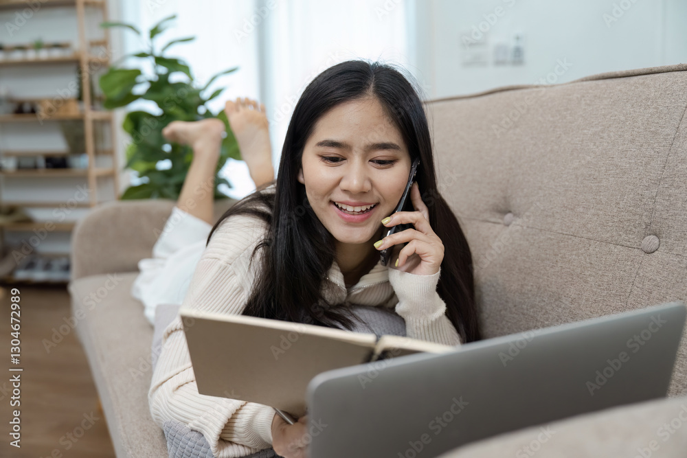 Young Asian woman chatting on the phone while studying from a laptop and reading a notebook at home. Young Asian woman chatting on the phone while studying from a laptop and reading a notebook at home.