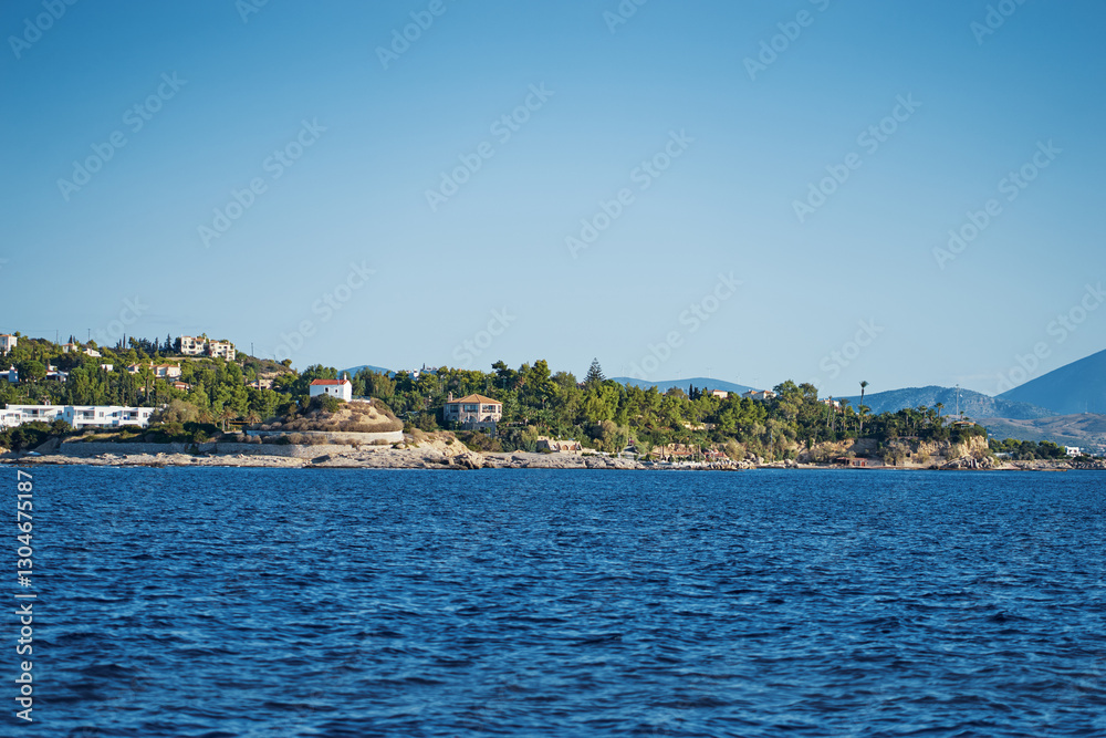 Naklejka premium A beautiful coastal view of Greece island showcasing vibrant blue waters, lush greenery, and idyllic residential areas under a clear blue sky, conveying serenity and natural beauty.