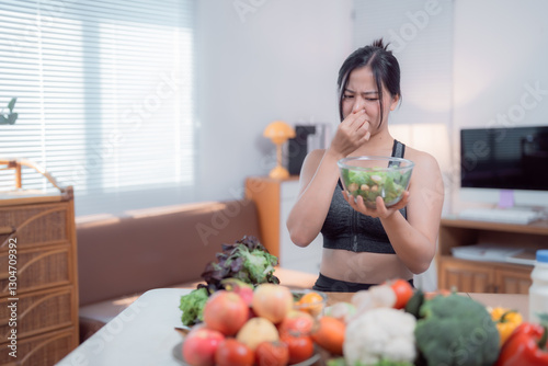 Young asian sportswoman is pinching her nose because of the bad smell coming from a bowl of spoiled salad in her kitchen, with many fresh vegetables and fruits on the table