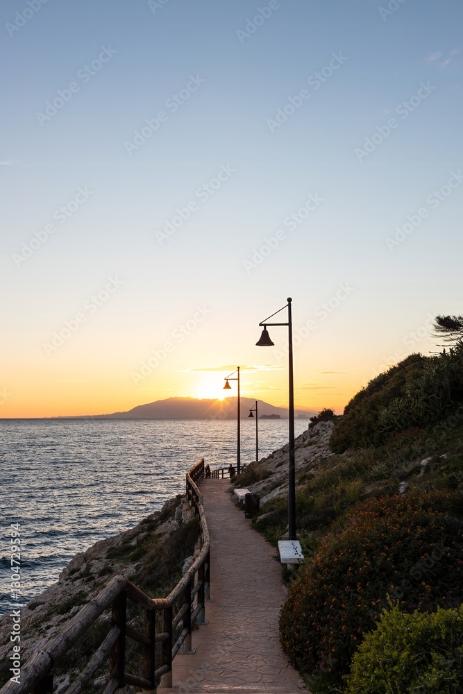 Obraz premium Evening promenade along a narrow pedestrian road along the sea at sunset. Sunlight illuminates the path. Street lights create a romantic atmosphere. Sunset walks along the sea.