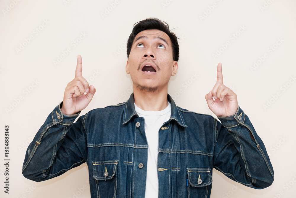 A Southeast Asian man wearing a denim jacket looking up with two finger pointing to up against a white background. This image depicts positivity, approval, and satisfaction A Southeast Asian man wearing a denim jacket looking up with two finger pointing to up against a white background. This image depicts positivity, approval, and satisfaction