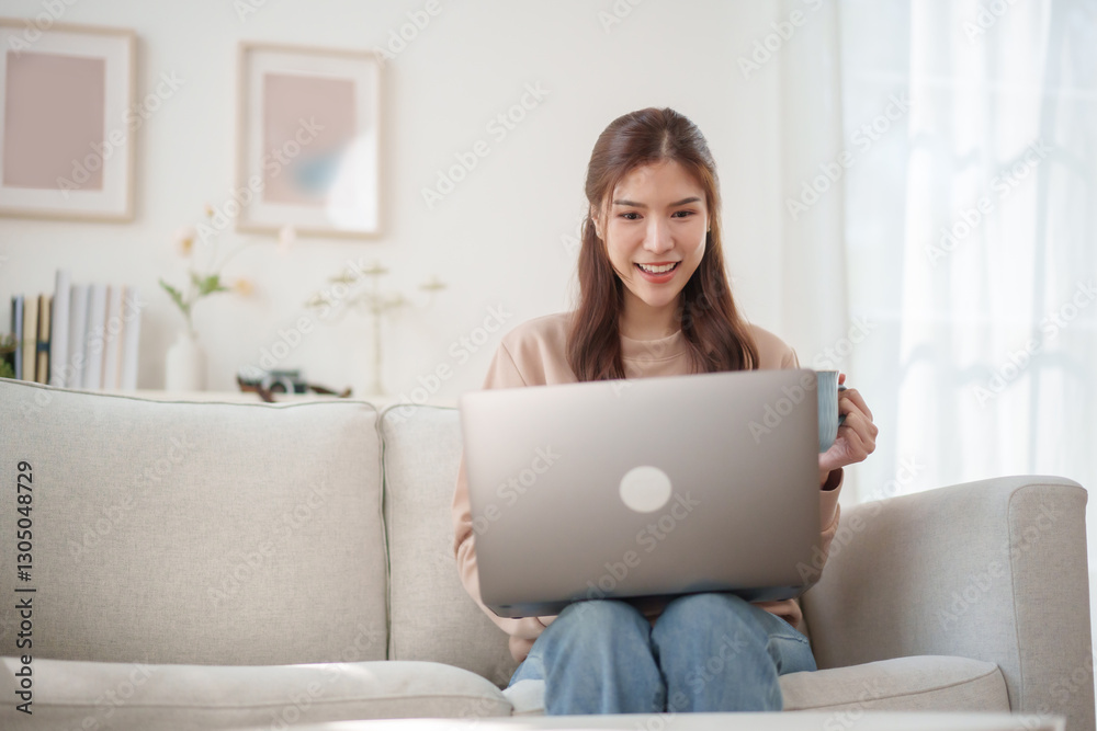 Beautiful Asian woman sits comfortably on a sofa, using laptop. smiling and interacts with her devices. Working at home. Beautiful Asian woman sits comfortably on a sofa, using laptop. smiling and interacts with her devices. Working at home.