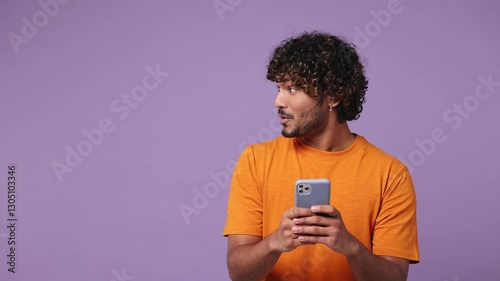 Young Indian man he wear orange t-shirt casual clothes hold use mobile cell phone look aside on workspace area copy space mock up isolated on plain pastel light purple background. Lifestyle concept