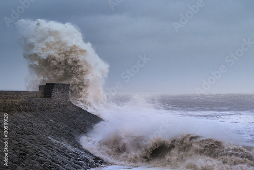 A huge winter storm hitting the coast of the UK. Giant waves crash into a breakwater, with a lighthouse. The sky is grey and moody