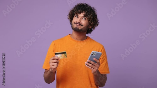 Young Indian man wear orange t-shirt casual clothes use mobile cell phone app hold credit bank card doing online shopping order delivery to home isolated on plain purple background. Lifestyle concept