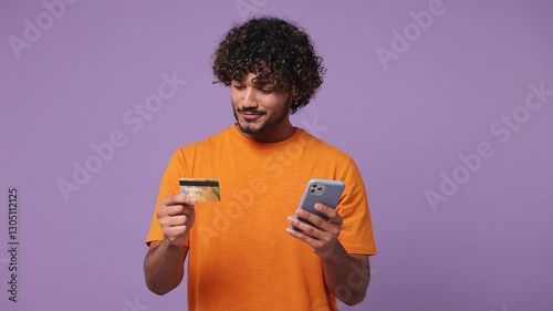 Young happy Indian man wear orange t-shirt casual clothes use mobile cell phone app hold credit bank card shopping online order delivery to home isolated on plain purple background. Lifestyle concept