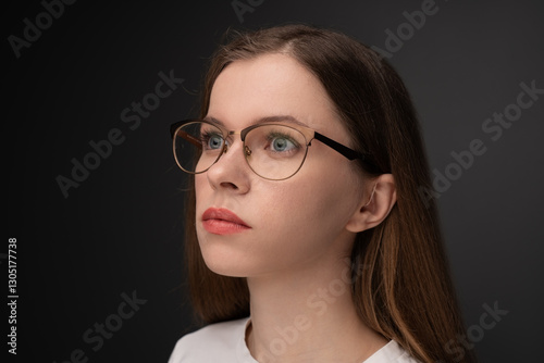 A young woman with long brown hair, glasses, and a receiver-in-canal (RIC) hearing aid looks thoughtfully into the distance. She wears a white shirt against a dark background, highlighting her face an