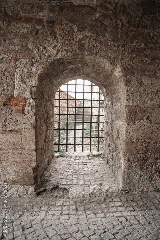Fototapeta premium Small grated doorway in the thick brick wall surrounding Kalemegdan Tower Castle. Belgrade, Serbia. Close-up of rusted iron portcullis corroded bars framed by cracked stone arch