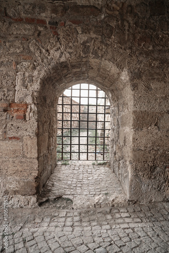 Small grated doorway in the thick brick wall surrounding Kalemegdan Tower Castle. Belgrade, Serbia. Close-up of rusted iron portcullis corroded bars framed by cracked stone arch