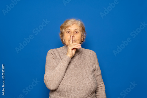 Caucasian woman about 70 years old wearing a dark beige sweater making the silence or shhh gesture keeping an important secret. Isolated on blue studio background.