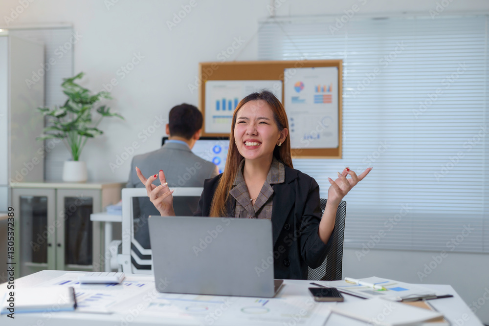Energetic Asian businesswoman celebrating success, raising hands while working on laptop in contemporary office space, colleague visible in background