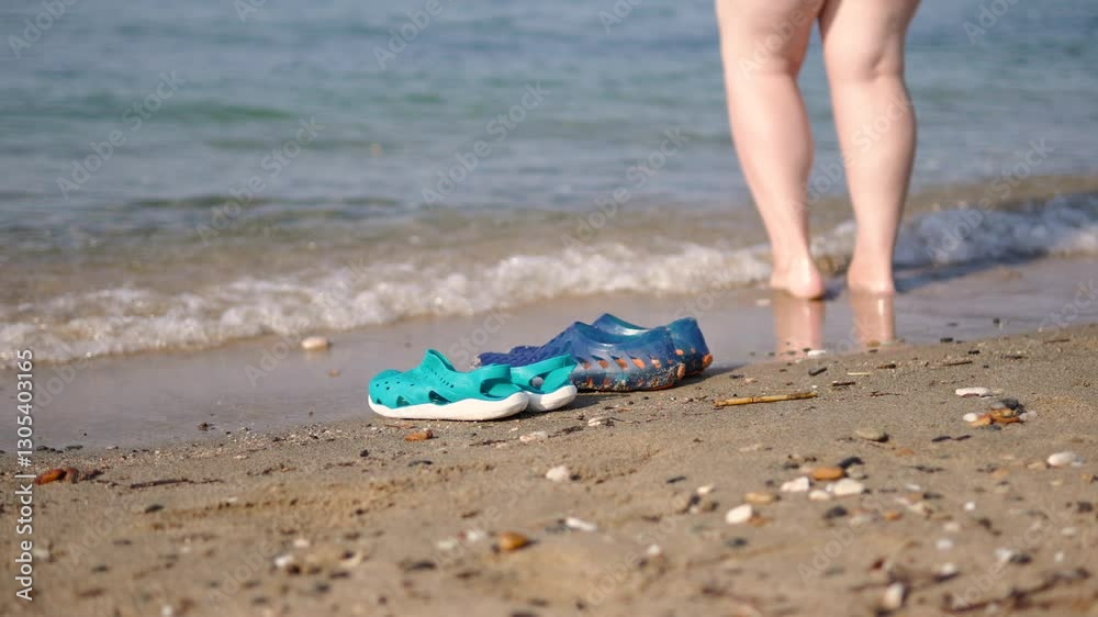 Beach waterproof shoes stand on sand near sea on sunny day. Light waves roll nearby and women bare feet walk on clear water. Protective footwear in form of aquasocks and clogs for swimming. Beach waterproof shoes stand on sand near sea on sunny day. Light waves roll nearby and women bare feet walk on clear water. Protective footwear in form of aquasocks and clogs for swimming.