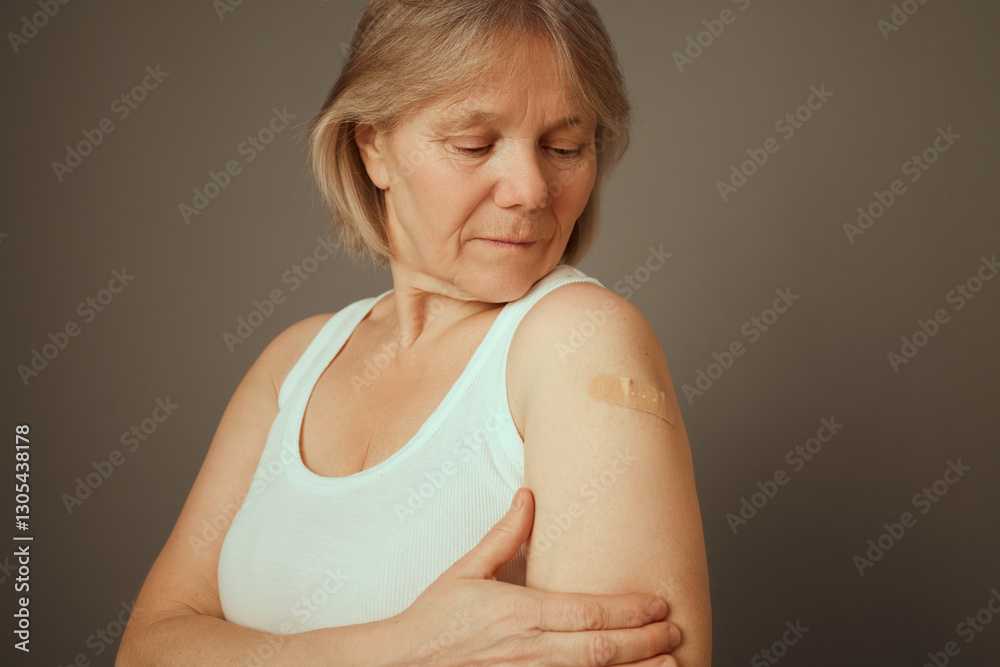 A middle-aged woman gently inspects her upper arm where a small bandage is placed after getting vaccinated, reflecting a moment of personal health awareness and care
