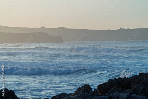 Coastal cliffs under a bright sun with powerful waves crashing on the shore. Surfers ride the swells, embracing the ocean's energy in this breathtaking seascape.
