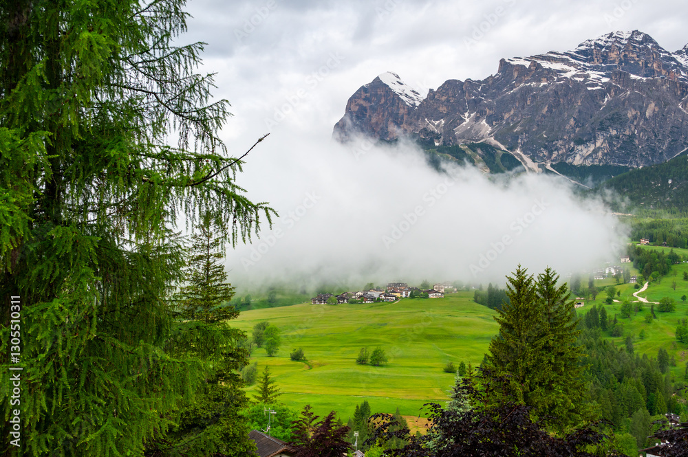 Scenic mountain village in the Dolomites (Dolomiti, Dolomiten), Italy. Cozy alpine houses on green slopes, towering peaks above, and clouds rolling through the valley below