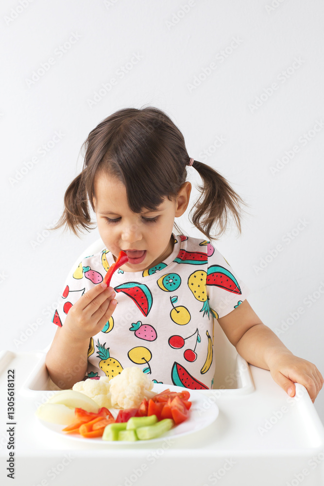 Little girl with pigtails sitting in high chair and eating slice of red bell pepper from plate of fresh vegetables, wearing colorful fruit-patterned t-shirt. healthy eating, childhood, nutrition Little girl with pigtails sitting in high chair and eating slice of red bell pepper from plate of fresh vegetables, wearing colorful fruit-patterned t-shirt. healthy eating, childhood, nutrition