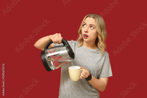 Thoughtful young woman pouring hot water from modern electric kettle into cup on red background
