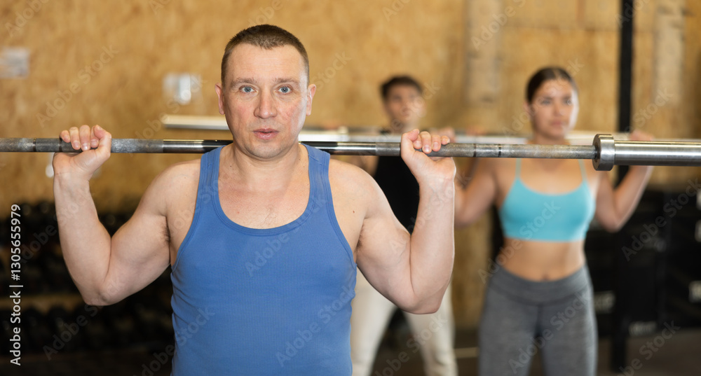 Focused middle-aged man in activewear exercising with barbell during group workout in gym health center. CrossFit healthy concept Focused middle-aged man in activewear exercising with barbell during group workout in gym health center. CrossFit healthy concept