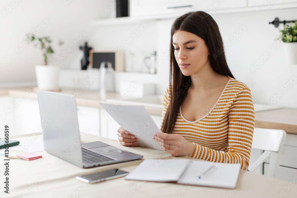 Focused young lady inspecting documents in front of her laptop at home in kitchen, woman doing paperwork Focused young lady inspecting documents in front of her laptop at home in kitchen, woman doing paperwork