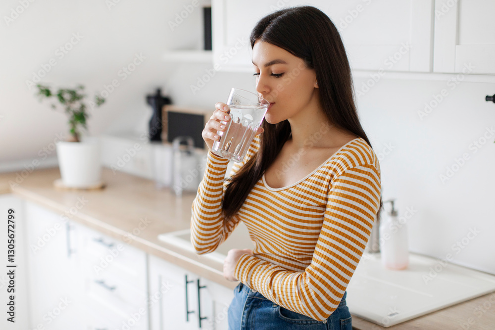 Young woman drinking clean mineral water from glass, standing in kitchen interior, free space. Healthy beverage, body hydration concept Young woman drinking clean mineral water from glass, standing in kitchen interior, free space. Healthy beverage, body hydration concept