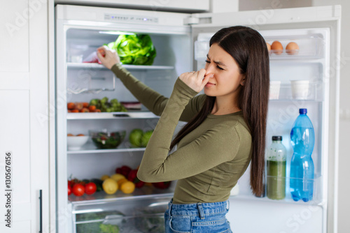 Lady holding nose cause of bad smell from refrigerator in kitchen, opening fridge, searching spoiled food