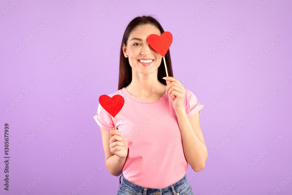 Playful Mood. Portrait of romantic girlfriend with red paper hearts in hands posing over purple studio background, happy affectionate woman covering one eye, looking and smiling at camera, free space Playful Mood. Portrait of romantic girlfriend with red paper hearts in hands posing over purple studio background, happy affectionate woman covering one eye, looking and smiling at camera, free space