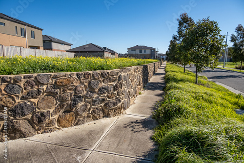 Foto A suburban street featuring a stone retaining wall running alongside a vacant plot and a concrete footpath with new modern homes in the background