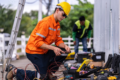 A skilled technician in a bright orange safety jacket and hard hat prepares electrical wiring with precision. Tools and open cases are spread in the background, showcasing a professional workspace.