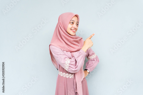 Asian Muslim young woman wearing headscarf veil hijab smiling cheerfully while looking and pointing to copy space beside her, standing on isolated gray background.
