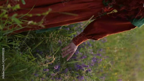 A shepherdess woman with red hair and a red and green dress walks on a field of flowers. Rural scene concept with a romantic peasant woman in a linen dress. Romance novel.