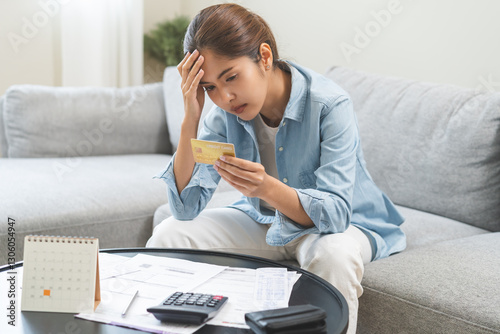 Debt and financial concept, stressed asian young woman have a headache while sitting on sofa calculate expenses, income, hand holding credit card for prepare payment or pay bills on table at home.