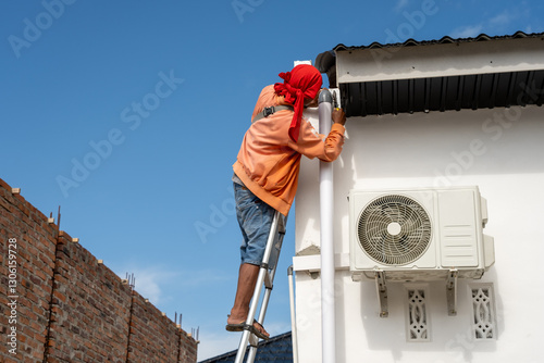 A construction worker in an orange hoodie and red headscarf installs a black metal awning on a white house while standing on a ladder. An air conditioning unit is mounted nearby
