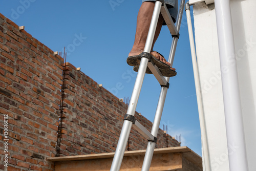 A construction worker stands on an aluminum ladder wearing flip-flops, working on a building under construction. A brick wall with exposed reinforcement bars is visible under a clear blue sky.
