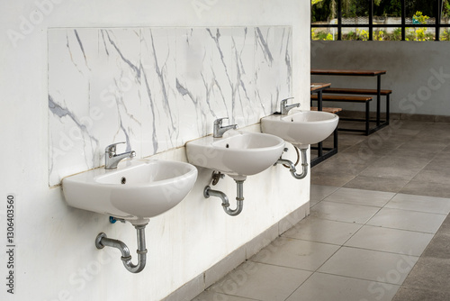Fototapeta Naklejka Na Ścianę i Meble -  A row of white ceramic sinks with chrome faucets, mounted on a marble-tiled wall in a public wash area. The background shows wooden benches and an open-air space with greenery.