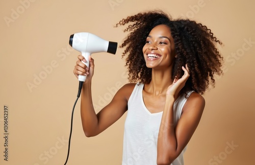 Smiling african american woman dries afro hair with blow dryer at home. Happy mixed race girl with healthy curly coiffure uses styling beauty products on beige background. Haircare concept.