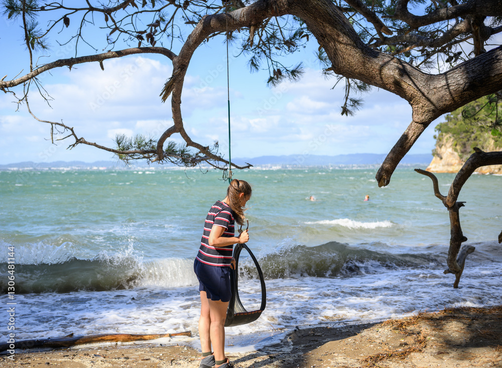 Woman checking the tyre swing on the beach. Unrecognizable people swing at Kendall Bay. Kauri Point Centennial Loop track. Auckland.