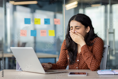 Boring monotonous everyday work. Woman at workplace sleepless yawning sitting at desk with laptop inside office. photo