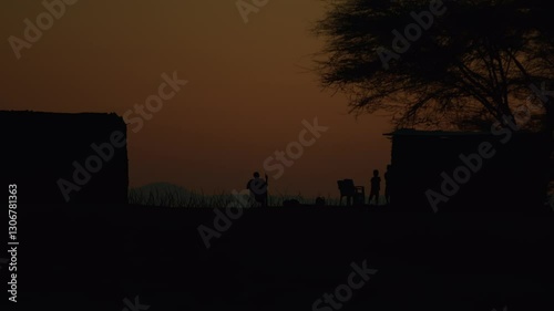 Kenya, Loima - September 9, 2023: Man resting in evening in chair near huts and children are playing near entrance to house. Houses in african settlements they are made of clay and covered with straw