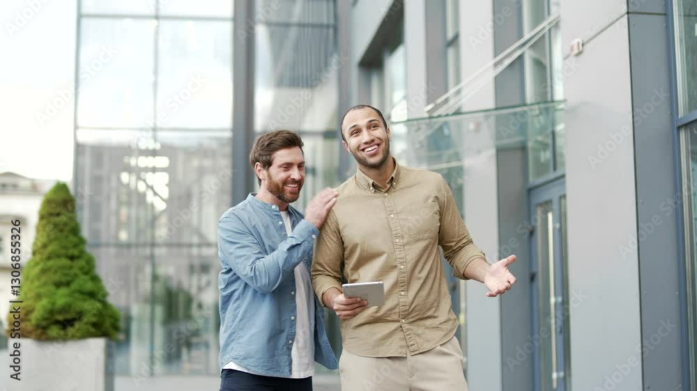 Two happy business colleagues have friendly conversation walking outside office building. Joyful worker praises friend for good results, achievements, congratulates shaking hands, smiling, rejoicing